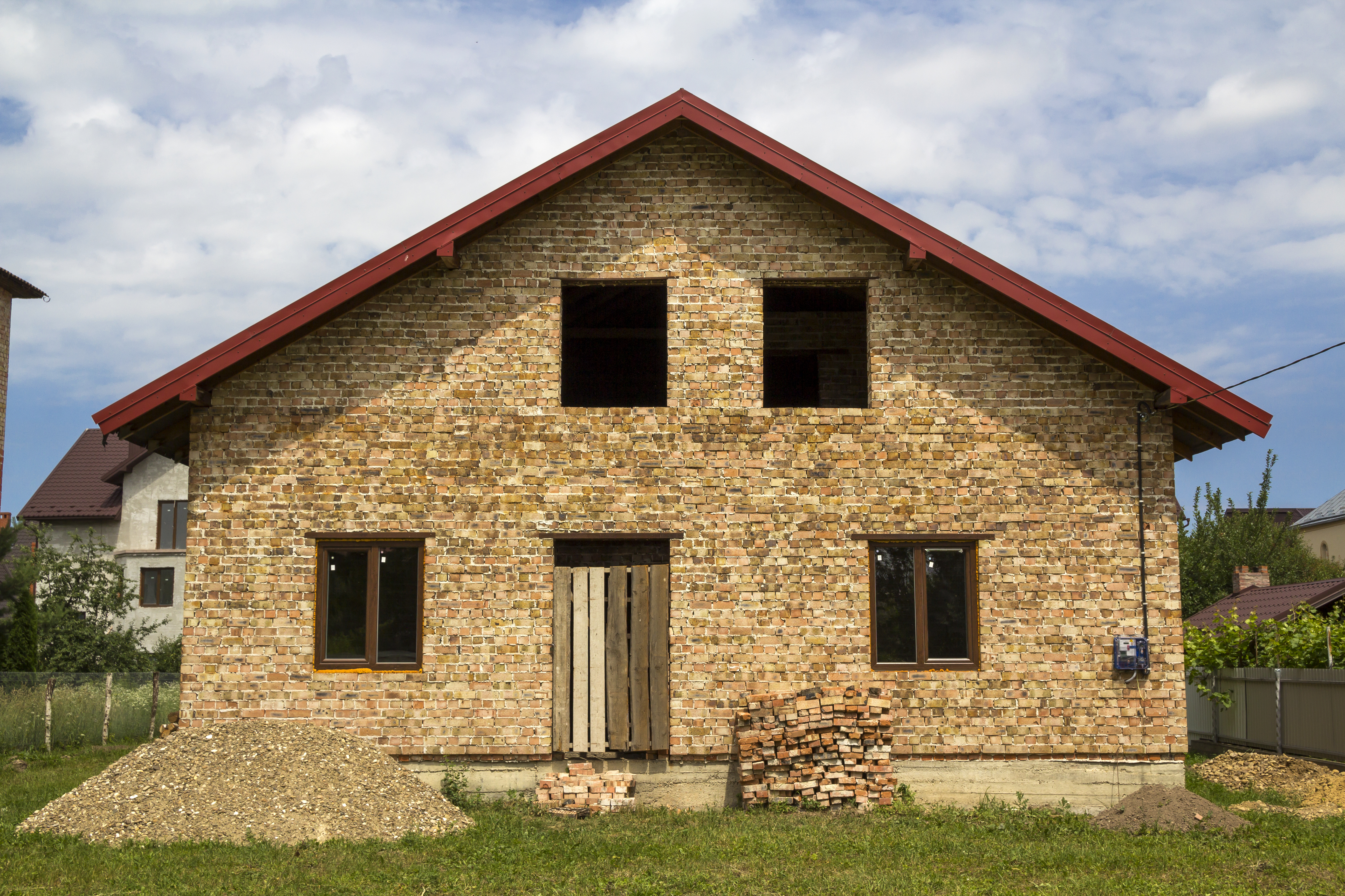 New not finished brick house with boarded door entrance, plastic windows on first floor, piles of brick and gravel on green grass on fenced land plot. Construction, investment and real property.